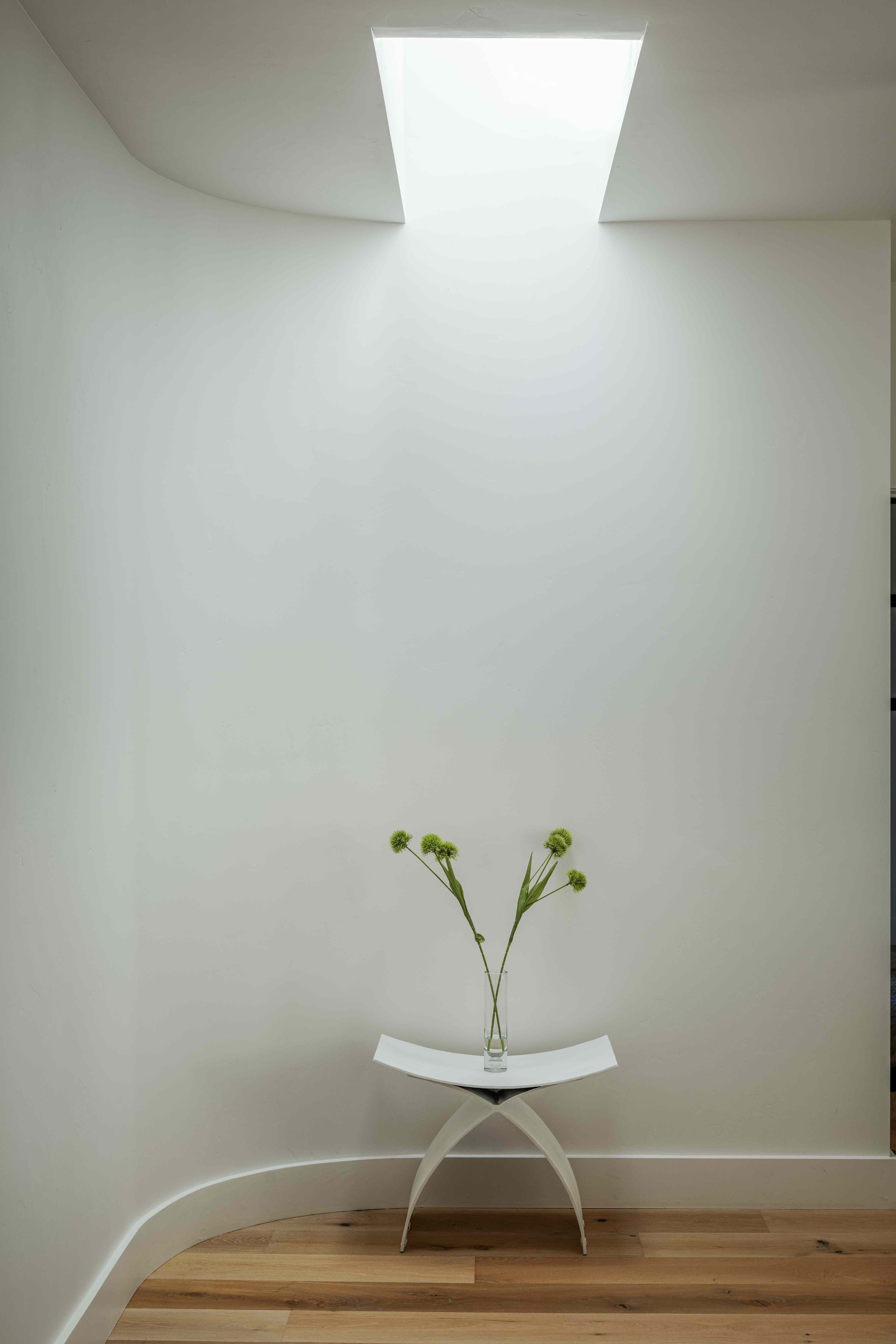 Minimalist stairwell with curved white walls, skylight and sculptural stool with green flowers